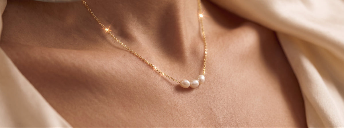 A close-up of a delicate gold chain necklace with three small white pearls, worn on a model's neck against a soft beige satin background.